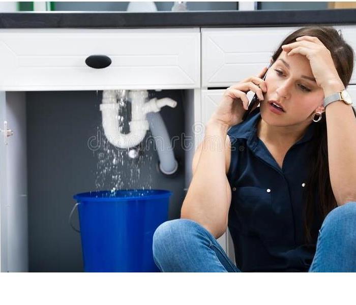 Upset homeowner on the phone with water gushing out of a busted pipe under the sink and using a bucket to catch the water. 